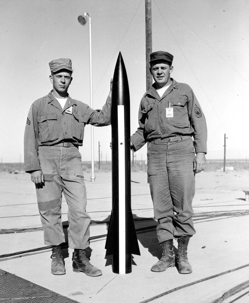 Two servicemen standing beside a rocket on a launch pad, with a clear sky in the background.